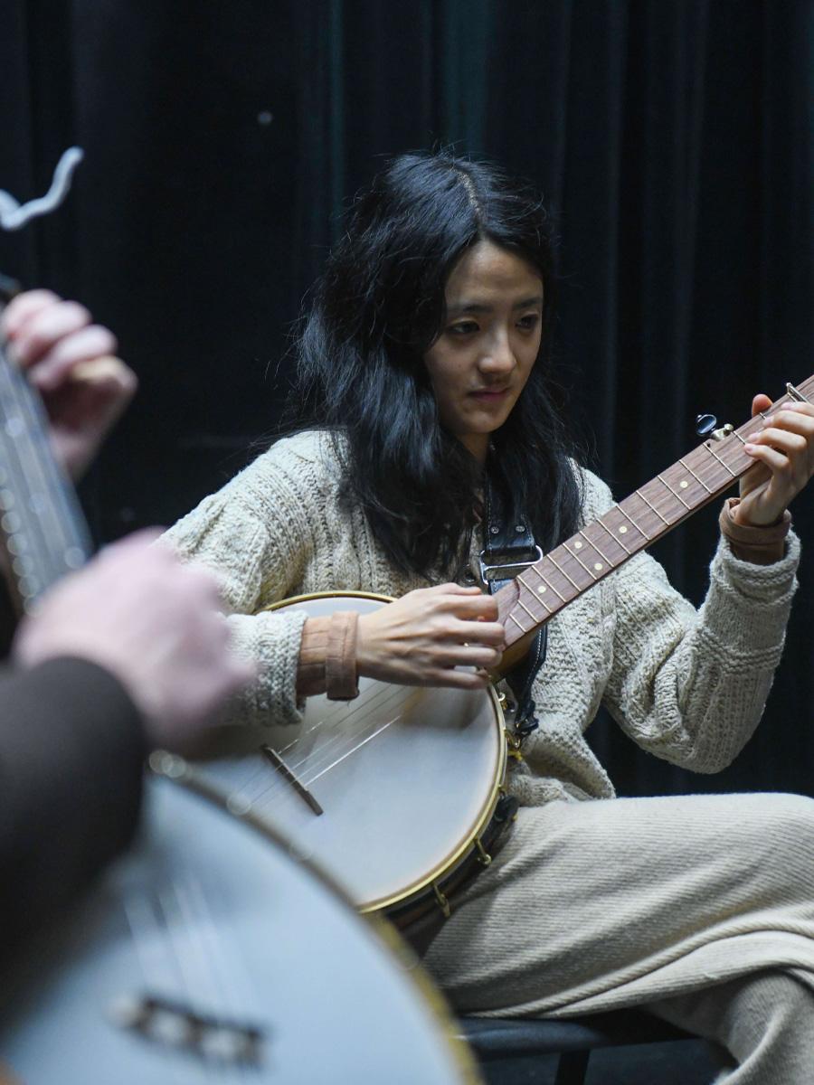 Banjo player playing in an ensemble with black hair and a white sweater.