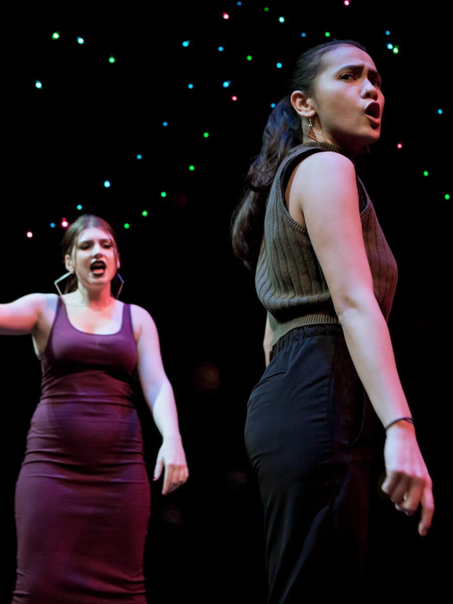Two women on a musical theater stage singing one left and one right in green tank top and black pants. 