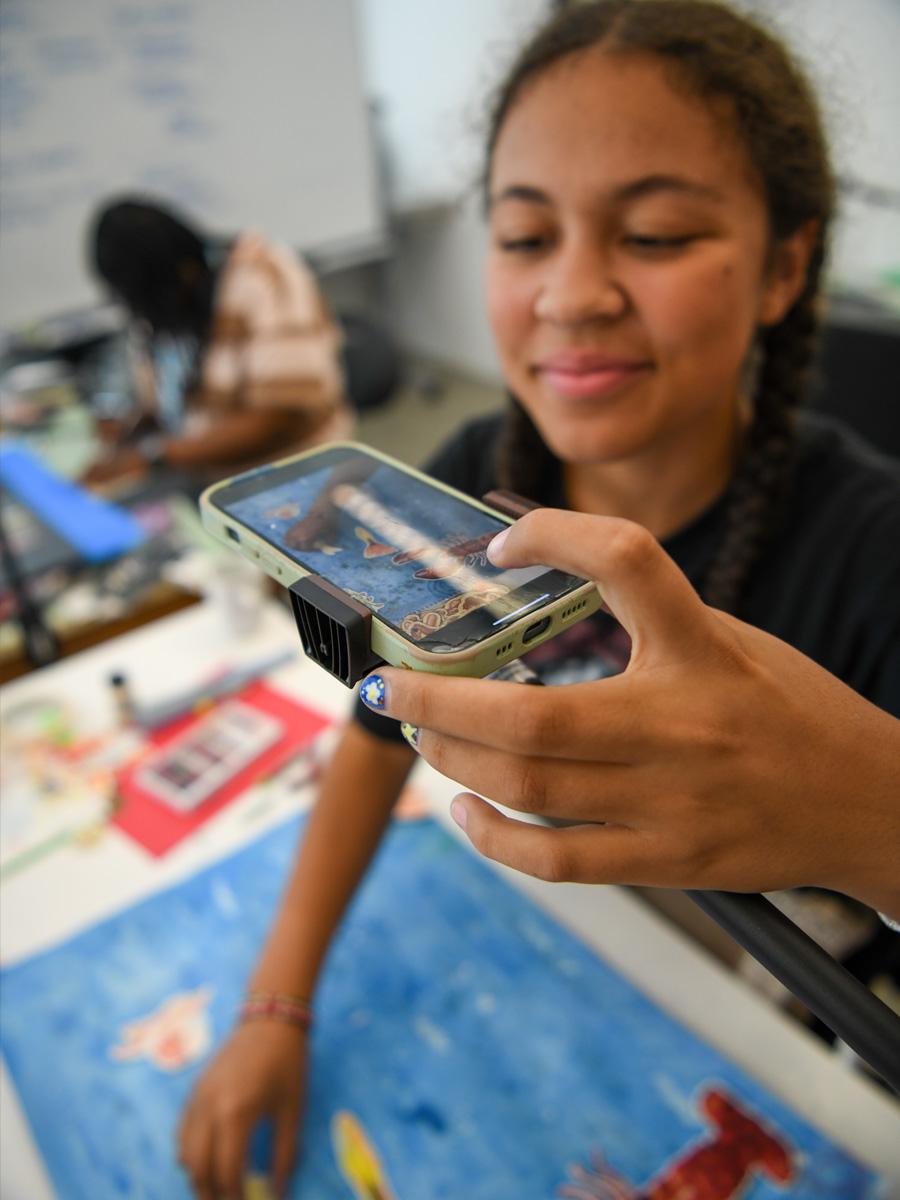 Student using a phone on holder to take photos of the stop motion scene on paper below in 2D. 