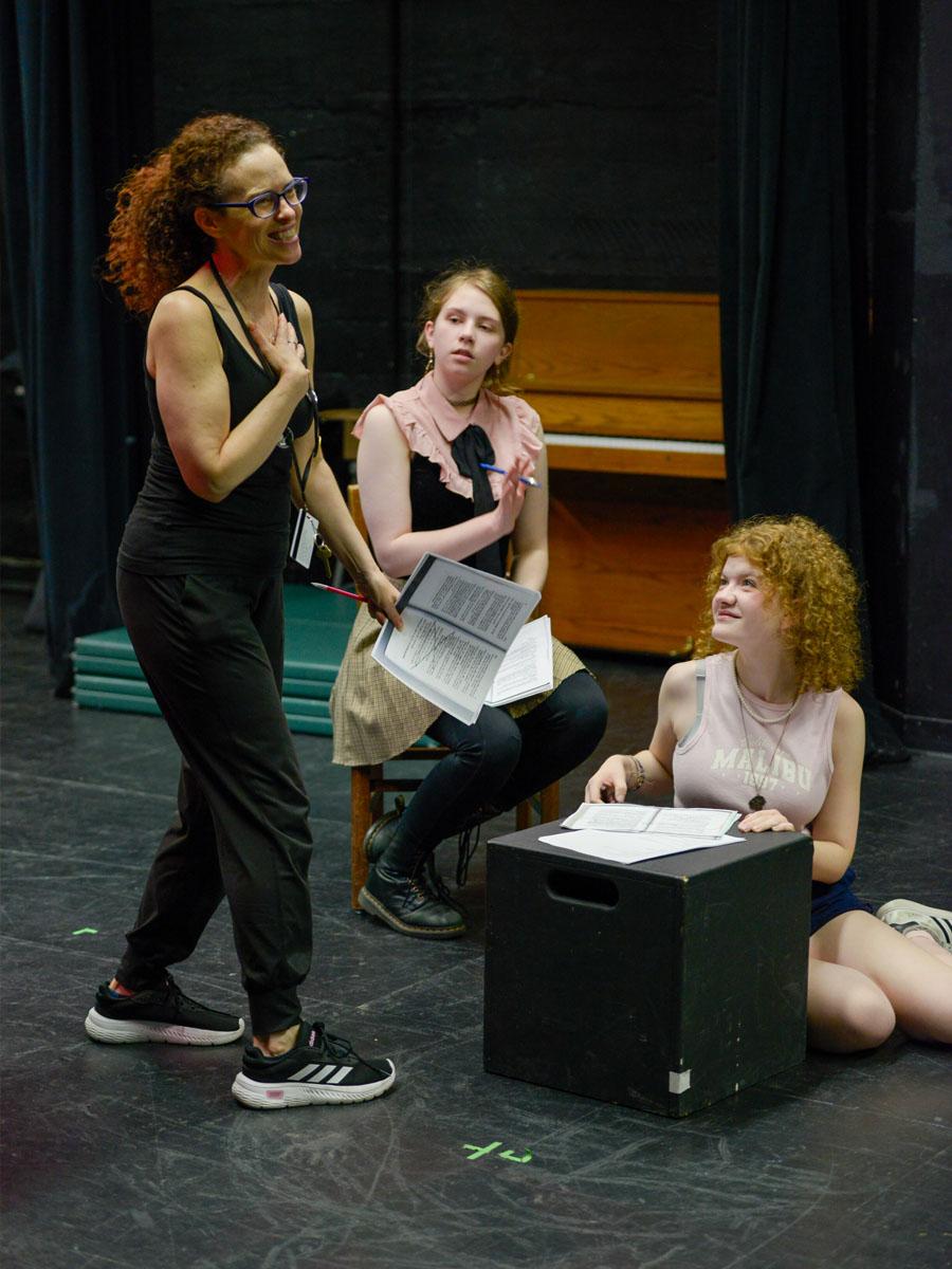 Two acting students sitting on acting blocks one with red hair and another on a chair in a black tank top next to the instructor