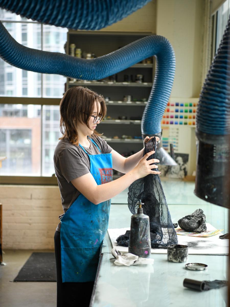 Analogue Printmaking Lab student working with blue tubes and glass desk with a student in a blue apron. 