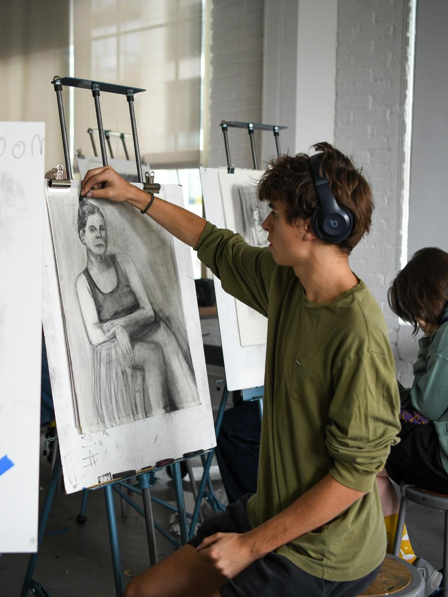 Student with headphones in green shirt charcoal drawing a model on paper. 