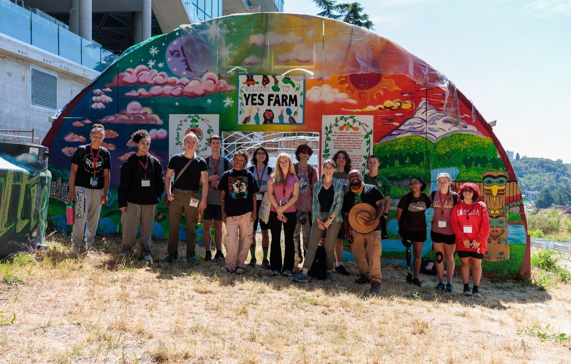 Group of students outside in front of a garden in the summer wearing colorful clothing. 