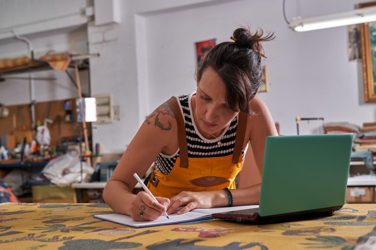 Artist writing and working with an apron on in front of a green laptop