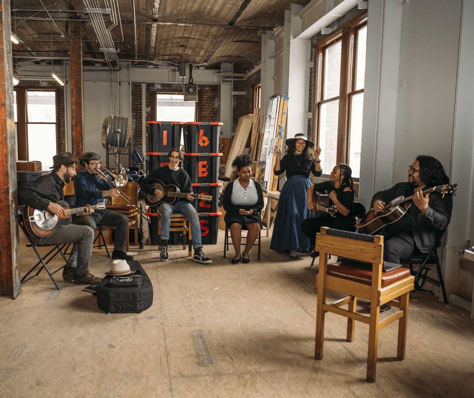 Seven musicians from The Rhapsody Project sitting in an old industrial-looking space playing music together. There are four people with guitars, a trombonist, and another two that look to be singers.
