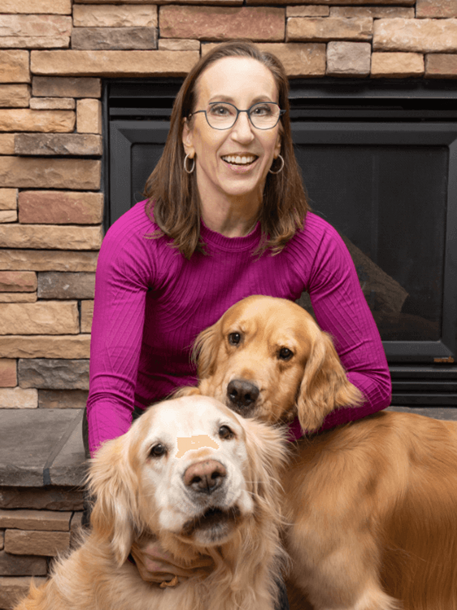 Woman wearing purple long[-sleeve shirt smiling with glasses and brown straight hair with her two golden retriever dogs.