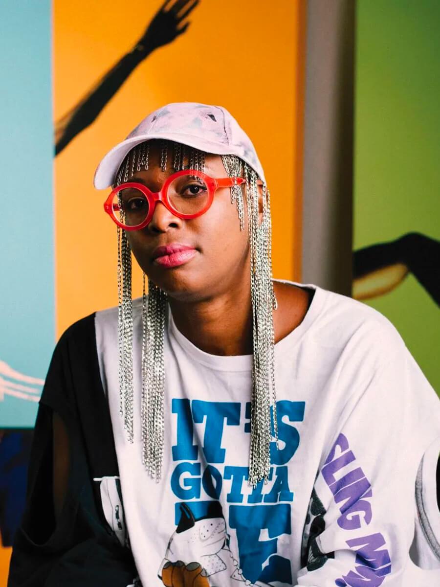 Dawn Okoro seated for a portrait wearing red framed glasses and a white baseball cap.