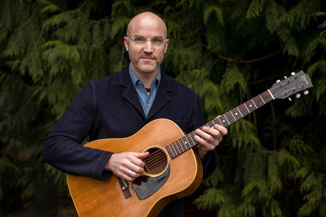 Eli West in a blue sweater holding his guitar in front of pine trees. 