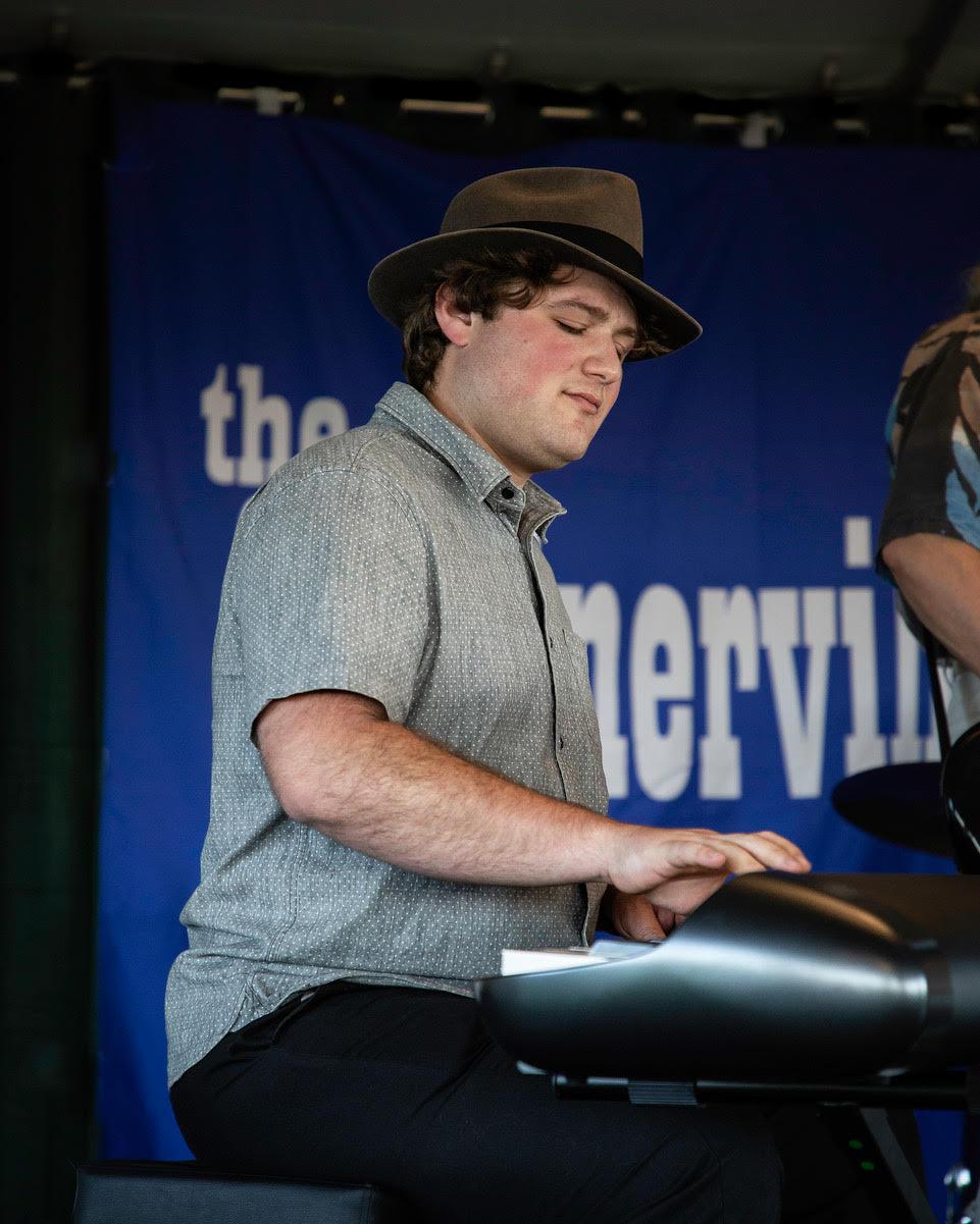 Tyler in a green shirt with a brown fedora hat on playing piano. 