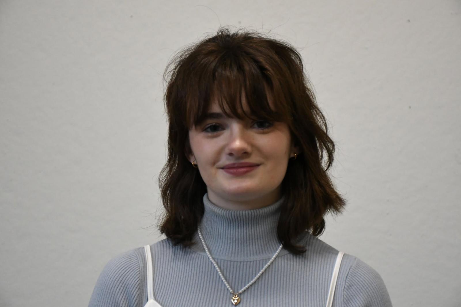 Abigail Roll in a sweater with a white tank top and brown hair. 