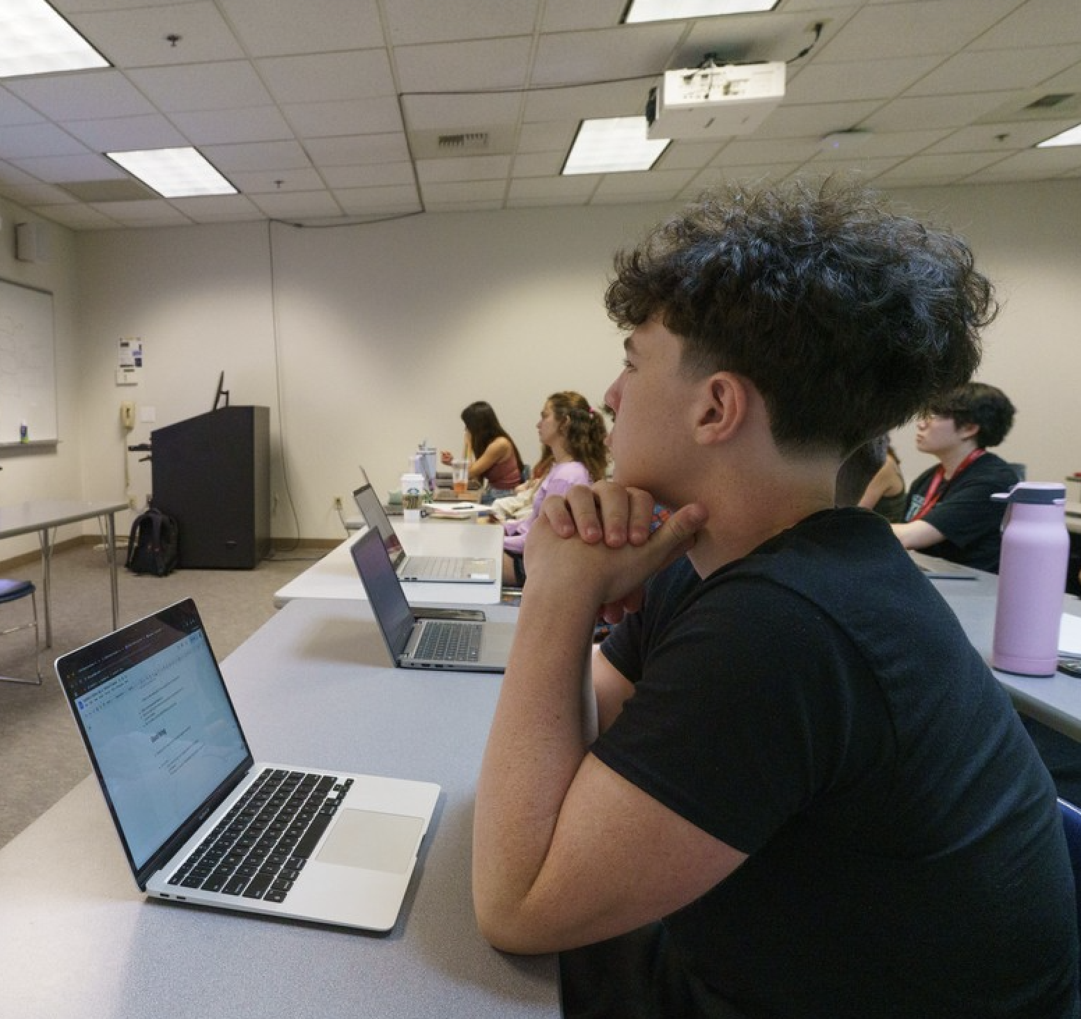 A student in a classroom in a black shirt and laptop watching a lecture. 